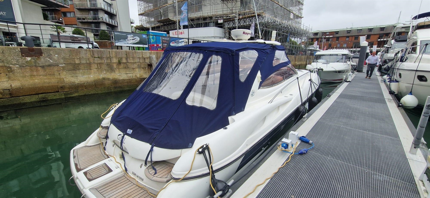 A white motorboat with a blue canopy is docked at a marina, with a man walking nearby on the boardwalk.